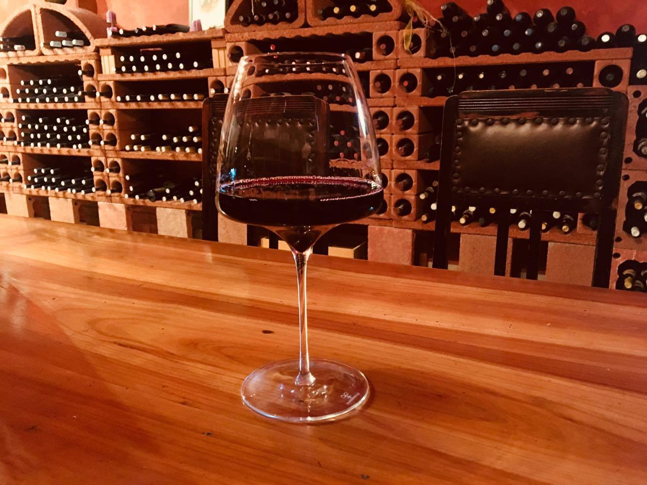 Ein Glas mit Rotweins steht auf einem Holztisch, dahinter zwei Stühle mit Lederbezug. Im Hintergrund ein Weinregal aus gebranntem Tonelementen in dem verschiedene Flaschen lagern.
